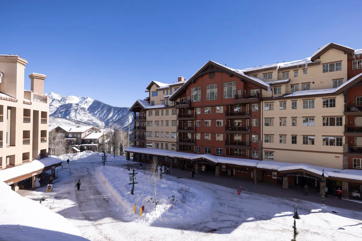 An exterior winter scene featuring a snow-covered landscape, mountain backdrop, and people enjoying themselves outside the resort buildings. The mood is festive and cozy, with hints of communal warmth and seasonal activities like skiing and outdoor dining.