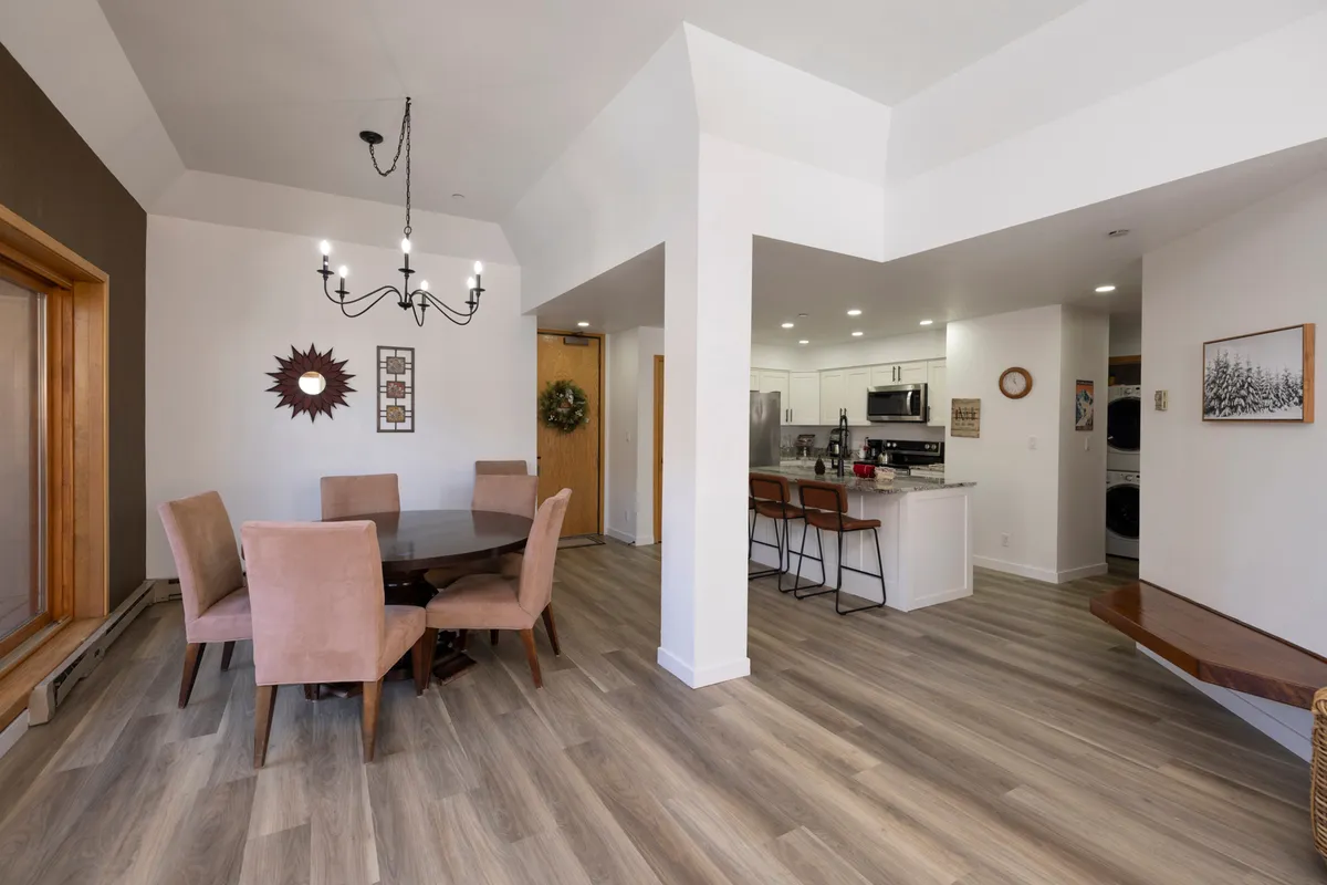 An open-plan view of the dining table with a glowing chandelier, adjacent to a modern kitchen area. Large windows offer additional natural light and mountain views, while the layout encourages effortless flow between cooking, dining, and socializing.
