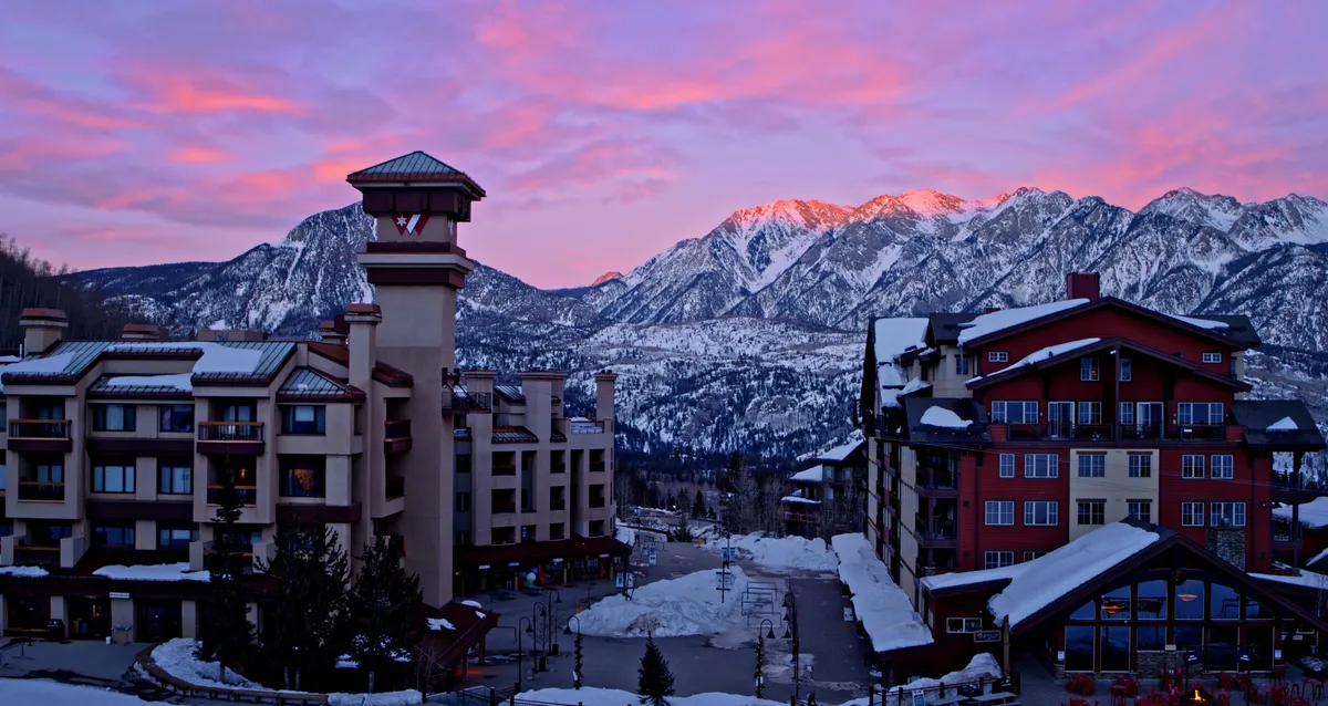This image displays the snow-covered mountains of Purgatory with a colorful sunset sky and rustic village architecture. The scene hints at various outdoor activities such as skiing, snowboarding, and mountain hiking, set against a peaceful evening backdrop.