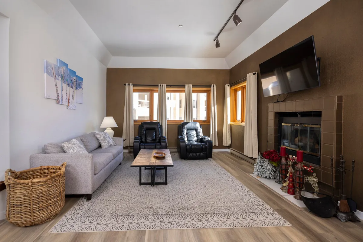 The image showcases a warm, inviting living area featuring a plush sofa, a stylish coffee table, and a stone fireplace surrounded by large windows that frame the mountain scenery. The setting exudes comfort and is perfect for relaxing, socializing, or enjoying a fireside evening after a day on the slopes.