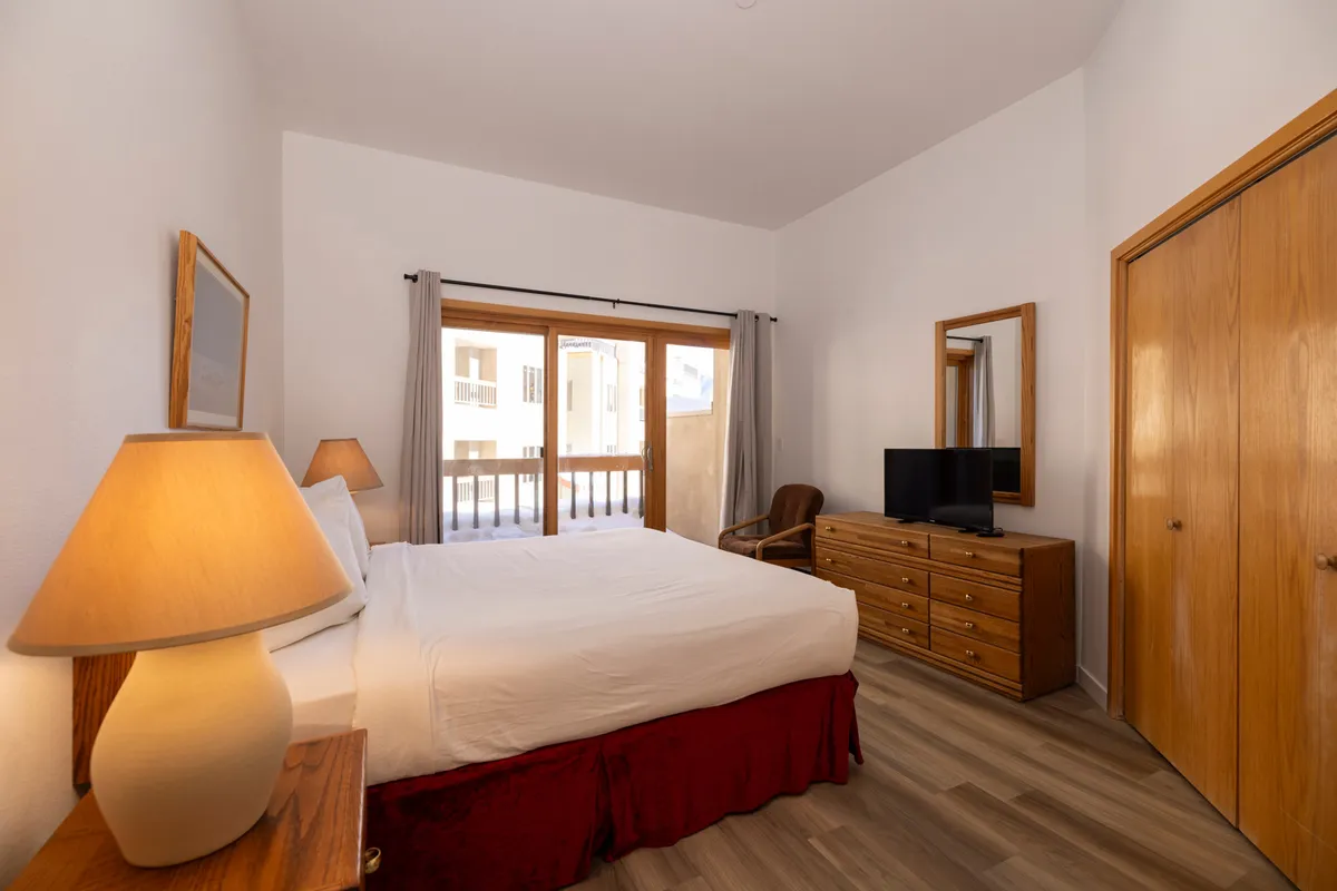 A warmly lit bedroom featuring a plush bed, cozy linens, a window that lets in natural light, a dresser, and a bedside lamp. The atmosphere is peaceful and inviting, perfect for restful sleep or a quiet evening of reading.