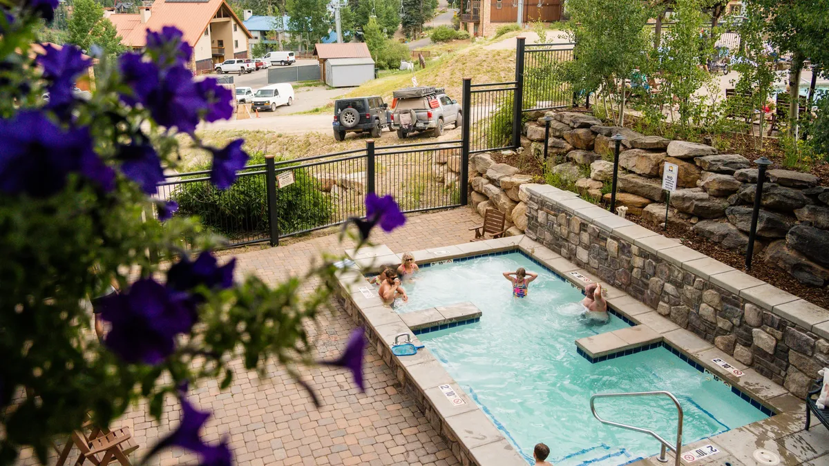 The image features a scenic outdoor hot tub and swimming pool surrounded by snowy landscapes and flowering plants, with guests relaxing and enjoying the water. The mountains are visible in the distance, and the mood is joyful and rejuvenating, suggesting après-ski relaxation.