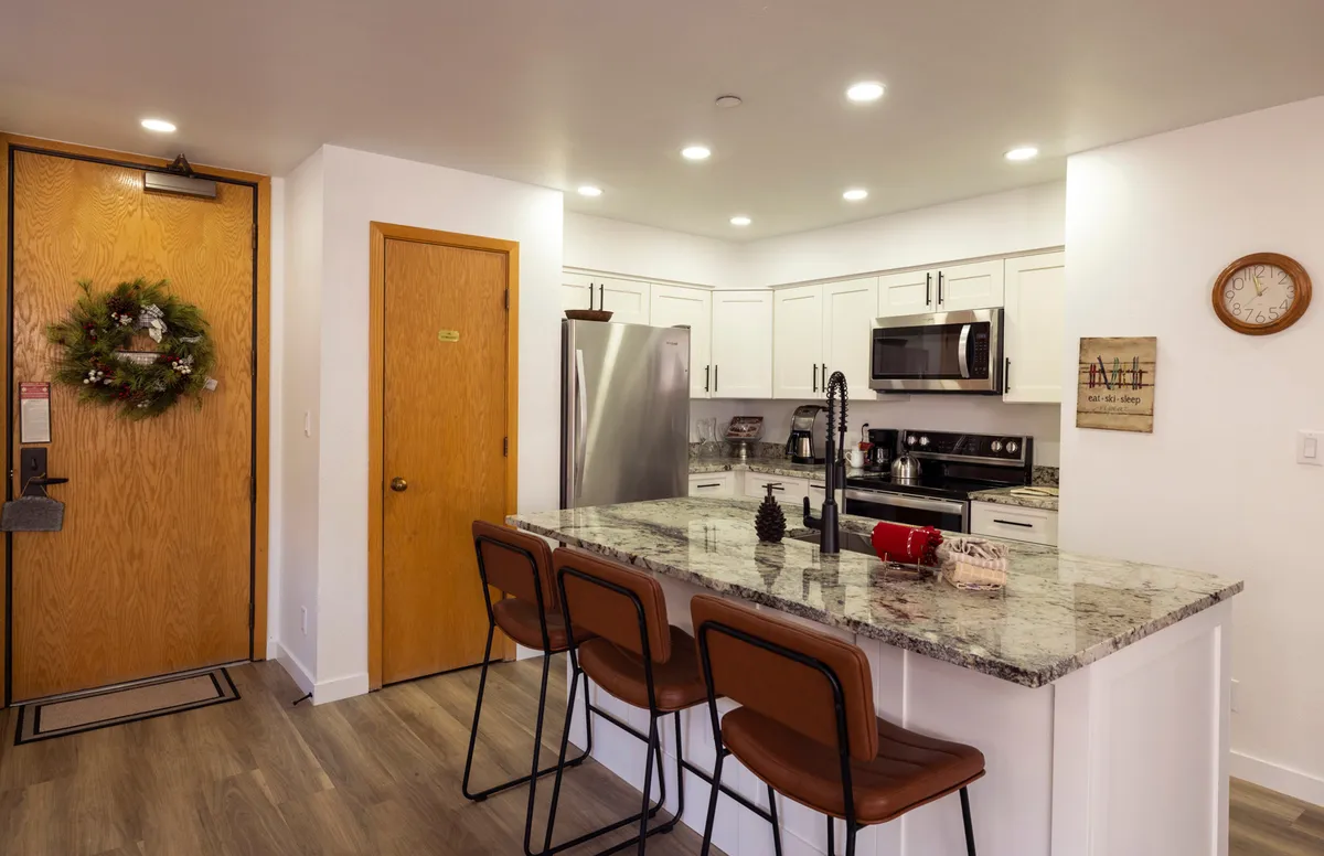 This image showcases the entryway and kitchen area, with a clear view of the entry door and a welcoming decorative wreath. The kitchen island with bar stools is visible, suggesting a space that bridges arrivals and departures, perfect for transitioning straight from the slopes to the heart of the home. The natural light and cozy décor evoke a sense of warmth and immediate access.