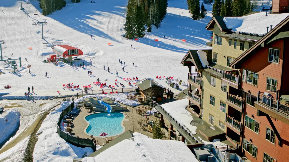 An exterior view of the lodge with a swimming pool set against the backdrop of ski slopes and a ski lift. The inviting blue pool stands out amidst the winter landscape, while the lodge's cozy exterior and mountain setting emphasize access to both aquatic leisure and outdoor adventure.